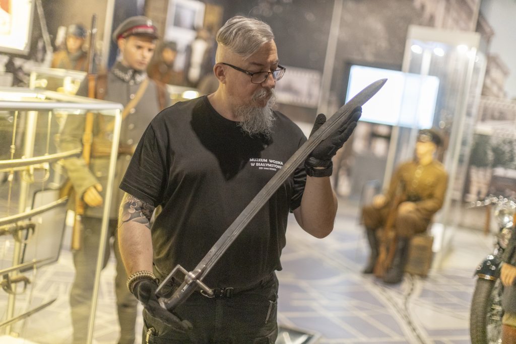 Fotografia. Mężczyzna prezentuje szable w dłoniach. W tle ekspozycja muzealna.