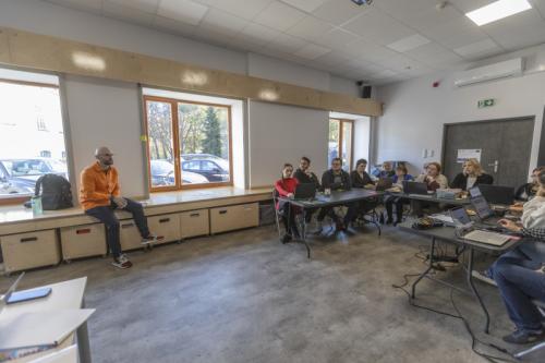 Sala edukacyjna. Po lewej stronie siedzi prowadzący warsztaty. Po prawej stronie uczestnicy warsztatów siedzący przy stołach ustawionych w literę U.